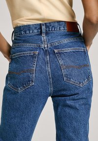 Υψηλής μέσης μπλε τζιν παντελόνι από denim, με δύο πίσω τσέπες με ανοιχτό ραφή και μια καφέ ετικέτα στη μέση.