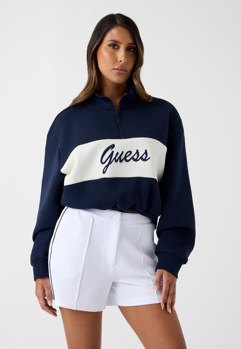 Femme debout, portant un sweat-shirt "Guess" bleu marine et blanc avec fermeture éclair et un short blanc à fines rayures noires sur les côtés, faisant face vers l’avant.