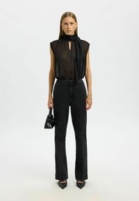 Haut noir transparent avec un nœud au col, associé à un jean noir taille haute à jambes droites. Le modèle tient un petit sac à main noir. Chaussures noires pointues.