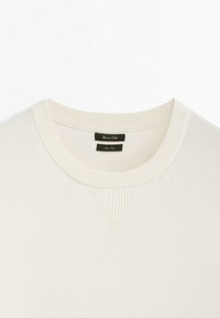 Pull en tricot couleur crème avec un col rond côtelé et un petit détail triangulaire côtelé sous le col.