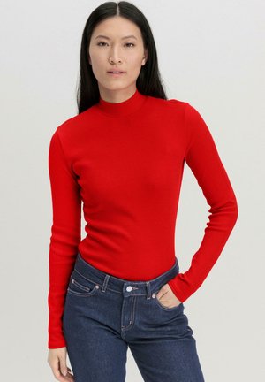 Rotes langärmliges Rollkragenoberteil aus geripptem Stoff, taillierte Passform, kombiniert mit dunkelblauen Denim-Jeans mit Knopfverschluss und Fronttaschen.