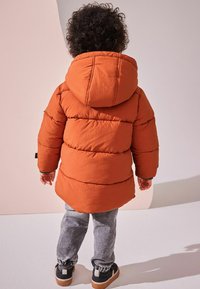 Oranžová péřová bunda s kapucí, s prošitým designem. Kombinována se šedými úzkými kalhotami a černými teniskami s lehkou podrážkou.