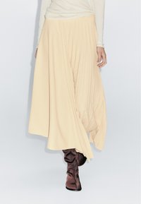 Beige asymmetrische rok met plooien, gecombineerd met donkerbruine slouchy laarzen, gedragen door een persoon met een lichtkleurige top met lange mouwen.