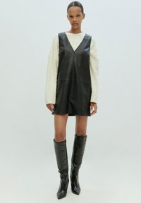 Schwarzes Lederkleid ohne Ärmel mit V-Ausschnitt und mittigem Reißverschluss, kombiniert mit einem strukturierten cremefarbenen Pullover und knielangen schwarzen spitzen Stiefeln.