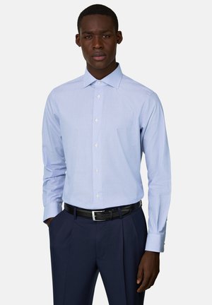 Mann trägt ein hellblau gestreiftes Hemd, in marineblaue Hose mit schwarzem Gürtel gesteckt, steht mit einer Hand in der Tasche.