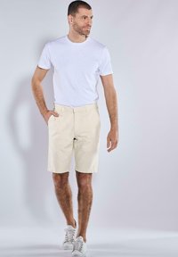White crew neck t-shirt, beige cotton shorts, and white sneakers. Model stands with one hand in pocket, displaying a casual outfit.
