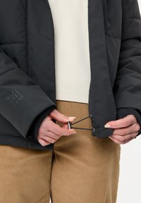 Veste matelassée noire à finition texturée, avec poignets élastiques et réglage par cordon à l'ourlet, portée avec un pantalon en velours côtelé beige.