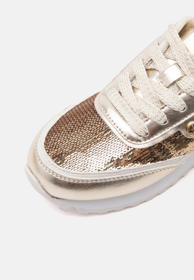 Κοντινή λήψη σε χρυσό sneaker με παγιέτες, λευκά και χρυσά κορδόνια και μεταλλικό χρυσό περίγραμμα σε λευκή σόλα.