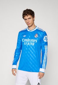 Man wearing blue Real Madrid goalkeeper jersey with long sleeves and white shorts, standing against plain background.