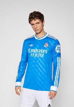 Uomo che indossa una maglia da portiere del Real Madrid blu con maniche lunghe e pantaloni bianchi, in piedi di fronte a uno sfondo neutro.