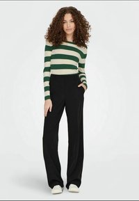 Striped sweater in cream and green with ribbed texture paired with high-waisted black trousers. Model stands with hands in pockets.