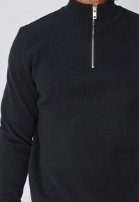 Pull noir à manches longues avec un col haut, doté d'une fermeture éclair en argent et d'un tissu en maille texturée. Design simple et ajusté.
