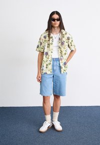 Kortærmet blomster skjorte i gul med blå og grønne mønstre, parret med lyseblå denim shorts og hvide sneakers med sorte accenter.