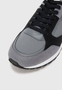 Grå sneakers laget av semsket skinn og syntetiske materialer, med en teksturert tåhette, svarte snørebånd, og hvit mellomsåle, detaljerte sømmer og gummisåle.