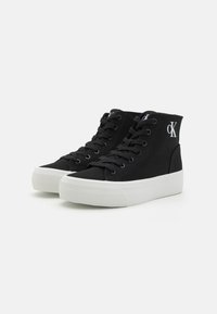Mustad mustad sneakers, millel musta kangast ja valge kummist tallaga. Omab musti paelu ja valget logo külgedel.