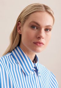 Camicia a righe blu e bianche con colletto classico e chiusura con bottoni, caratterizzata da una trama di cotone liscia e motivi verticali uniformi.
