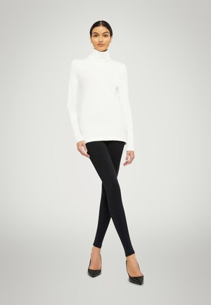 Maglione bianco a collo alto abbinato a leggings neri; la modella posa con le mani sui fianchi, indossando tacchi a punta neri su uno sfondo chiaro.