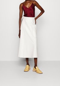 Witte knielange wikkelrok met een zak aan de voorkant en zichtbare stiksels, gecombineerd met een bordeaux satijnen cami-top en gele instap schoenen.