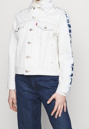 Person wearing white denim jacket with tie-dye blue patterns on sleeves, buttoned front, chest pockets, and dark blue jeans.