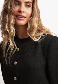 Mustmust tekstuuriga kardigan ümarate krae ja kullakarva nuppudega esiosas. Mudelil on pehmed, lainelised juuksed.