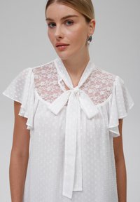 Hvit bluse med pufferm, blonder i halsen og en knyttet sløyfe ved kragen. Stoffet har et teksturert prikkemønster.