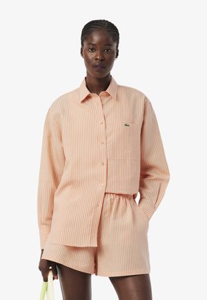 Femme portant une chemise à manches longues à rayures verticales pêche et blanches ainsi qu'un short assorti, tenant un sac à main jaune clair dans une main.