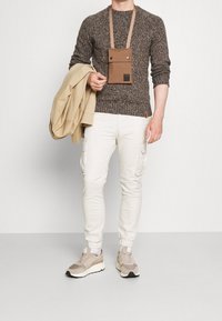 Brauner Pullover mit Struktur, beiger Cargohose mit Seitentaschen, graue und weiße Sneakers und eine hellbeige Jacke, die über dem Arm getragen wird. Braune Umhängetasche.