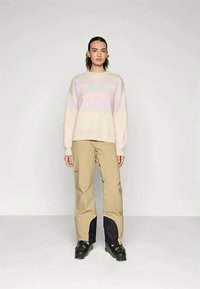 Ανοιχτό μπεζ πουλόβερ πλεκτό με παστέλ ροζ γεωμετρικό σχέδιο, συνδυασμένο με καμηλό cargo παντελόνι που έχει μαύρες λεπτομέρειες και τσέπες.
