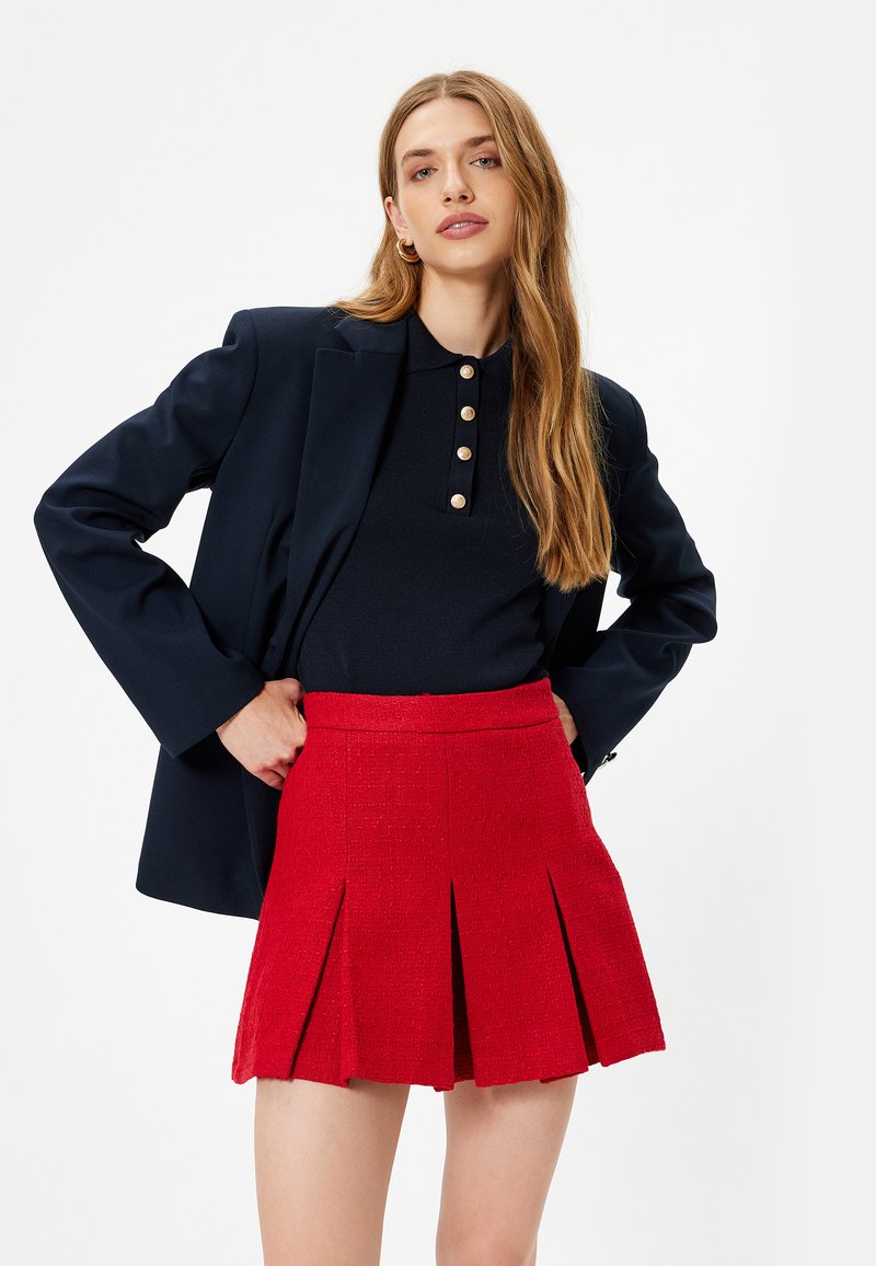 Rød plisseret nederdel lavet af tekstureret stof, parret med en sort polo med dekorative knapper, lagdelt under en mørk blazer.