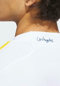 Vit sporttröja med gula detaljer, med en ribbad krage och "Los Angeles" broderat på baksidan av nacken i blått.