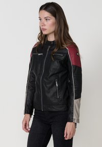 Giacca in pelle nera con spalle trapuntate rosse, dettagli in argento, tasche con zip e colletto alto. Presenti dettagli testurizzati sulle maniche.