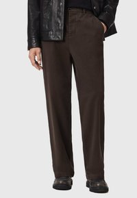 Bruine, rechte broek gemaakt van gladde stof. Voorzien van zijzakken en een knoopsluiting, gecombineerd met zwarte schoenen en een leren jas.