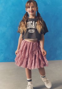 Graues cropped T-Shirt mit dem Schriftzug "CHICAGO USA", geschichteter rosa Tüllrock, weiße Sneakers und schwarz-weiße gestreifte Socken, vor einem blauen Hintergrund.
