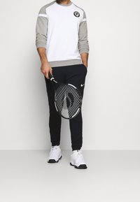 Maglietta a maniche lunghe bianca e grigia con scollatura rotonda, pantaloni sportivi neri e scarpe da tennis bianche, mentre tiene una racchetta da tennis nera. Logo Nike in vista.