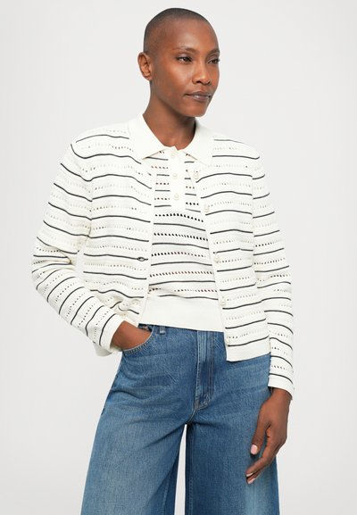 Femme aux cheveux courts portant un cardigan en tricot rayé noir et blanc sur un polo assorti et un jean bleu taille haute, la main dans la poche.