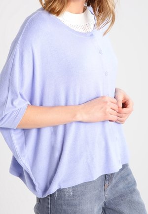 Frau trägt ein lockeres, hellviolettes Hemd über einem weißen Shirt und ausgewaschene blaue Jeans, von den Schultern bis zu den Hüften seitlich abgebildet.