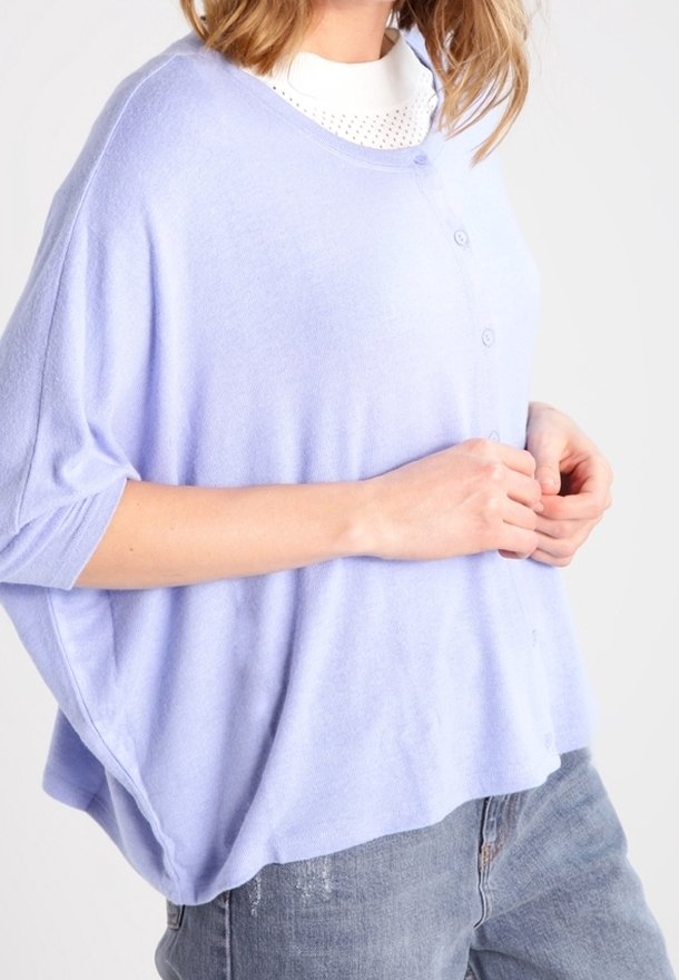 Femme portant un haut boutonné ample violet clair sur un t-shirt blanc et un jean bleu délavé, montrée des épaules aux hanches, de profil.