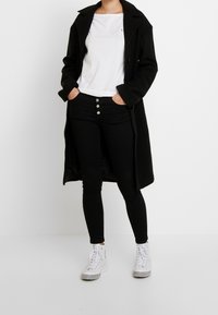 Person in einem schwarzen langem Mantel, einem weißen T-Shirt, schwarzen hoch taillierten Hosen mit Knöpfen und weißen High-Top Sneakers vor einem weißen Hintergrund.