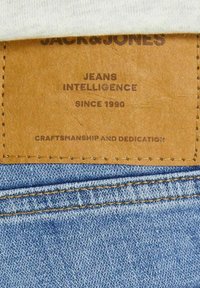 Light blue denim fabric with a brown leather patch labeled "JACK & JONES" and "JEANS INTELLIGENCE." Includes decorative stitching.