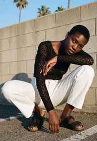 Vrouw met kort haar hurkt, gekleed in een zwart langärmelig shirt met luipaardprint, witte broek en zwarte sandalen tegen een muur van betonblokken.