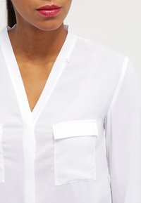 Vit, lätt blus med V-ringning, långa ärmar och två framfickor. Har en mjuk textur och subtila knapprundel.