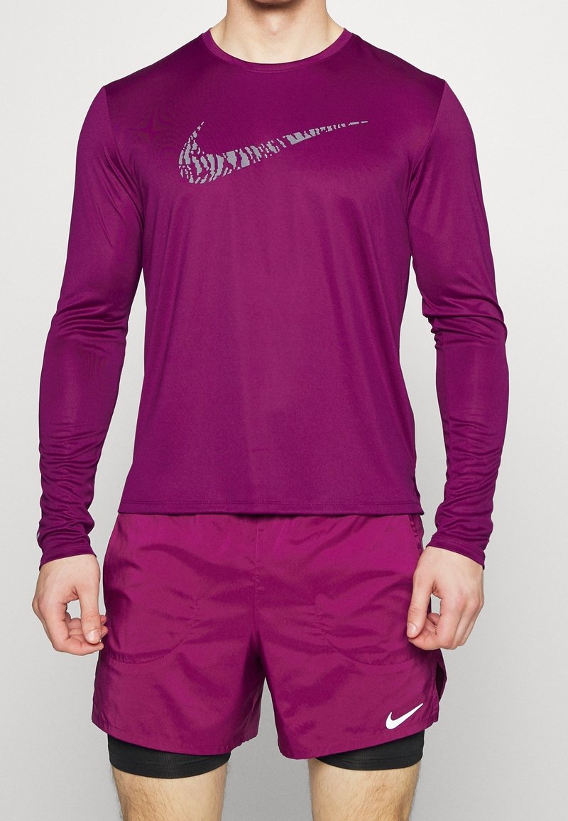Langärmliges lila Sportshirt mit reflektierendem Nike-Logo, kombiniert mit lila Shorts und schwarzen Unterhosen, hergestellt aus leichtem, dehnbarem Material.
