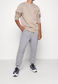 Person in beigem Nike-Kapuzenpullover, grauen Jogginghosen und schwarz-weißen Sneakern steht mit einer Hand in der Tasche vor einem weißen Hintergrund.