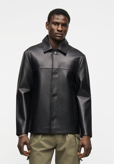 Άνδρας που φοράει μαύρο δερμάτινο μπουφάν με κουμπιά και γιακά, και ελιάς-πράσινο παντελόνι, στέκεται μπροστά σε ένα απλό φωτεινό φόντο.