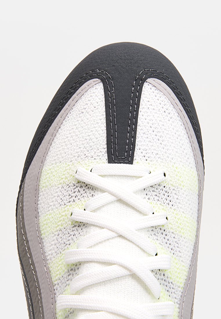 Scarpa sportiva bianca e grigia con tomaia in maglia testurizzata, punta in pelle nera e lacci bianchi. Presenta dettagli a strisce gialle.