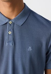 Polo bleu texturé avec deux boutons et un logo brodé "M OP" sur le côté gauche de la poitrine, porté par une personne.
