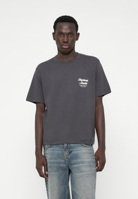 T-shirt cinza em algodão com decote redondo e mangas curtas, apresentando um gráfico de texto branco no lado esquerdo do peito. Combinado com jeans denim azul claro.