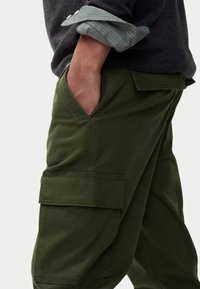 Groene cargobroek van duurzaam materiaal, met een zijvak voor cargo, diepe zijzakken en een relaxte pasvorm. Opgerolde mouwen van een geruite overhemd zichtbaar.