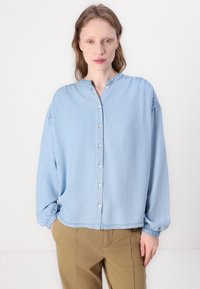 Lys blå, knapp-opp bluse laget av denim med kinesisk krage, lange ermer og elastiske mansjetter. Subtil samling ved skuldersømmer.