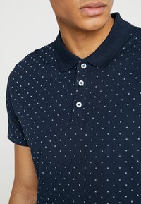 Námořnická polo košile vyrobená z bavlny, s límečkem a knoflíky. Má vzorovaný design s malými bílými diamantovými tvary.